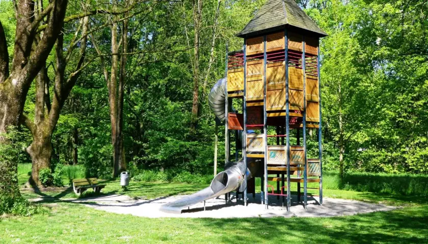 Ein hoher Holzturm mit Rutschen bildet das Zentrum eines abenteuerspielplatzes in Stuttgart. Umgeben von grünen Bäumen lädt er zum Spielen ein.