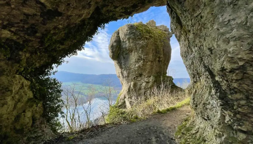 Blick durch eine Felsöffnung auf einen markanten Felsen mit Moosbewuchs. Ein Pfad führt entlang der Felsen. Eine tolle hohle Felsen Tour.