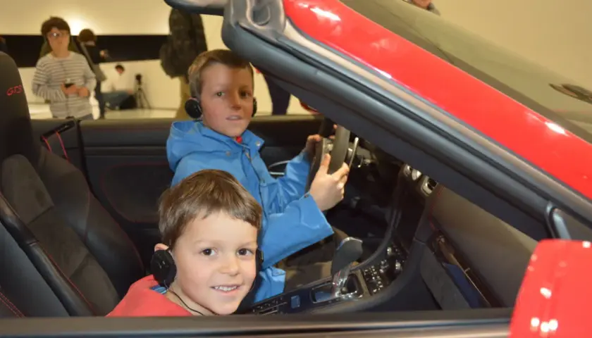 Zwei Jungen mit Kopfhörern sitzen in einem roten Porsche im Porsche Museum.