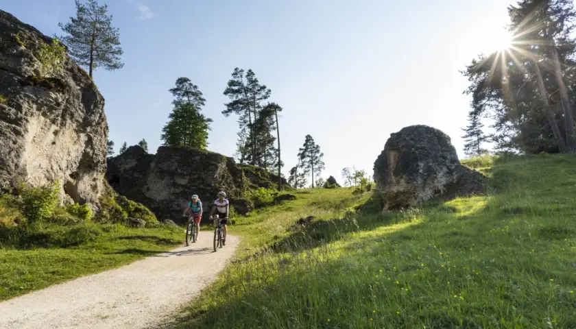 Zwei Radfahrer genießen eine Tour durch eine felsige Landschaft. Sie fahren auf einem Schotterweg, flankiert von grünen Wiesen und imposanten Felsen. Ein perfekter Tag, um in Heubach zu wandern.