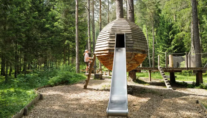 Ein Mädchen klettert auf einem Baumstamm an einem Waldspielplatz. Eine runde Holzhütte mit Rutsche steht im Mittelpunkt.
