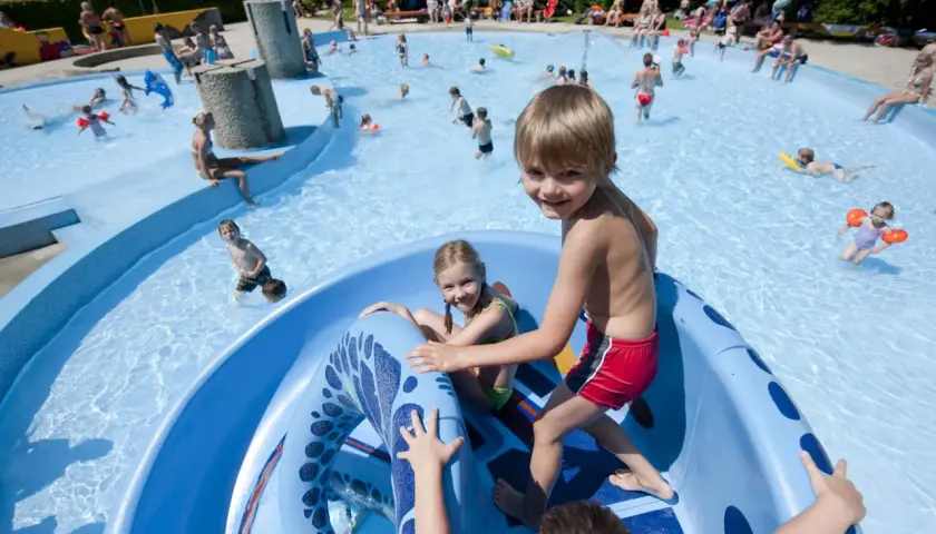 Fröhliche Kinder planschen und rutschen im Freibad. Ein Junge grinst in die Kamera, während ein Mädchen auf der blauen Rutsche sitzt. Ein perfekter Tag im bayerischen Wald mit Kindern.