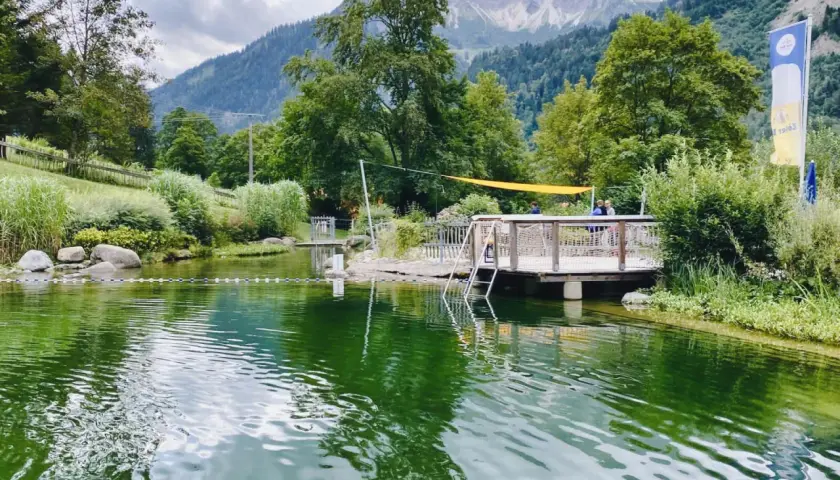 Ein idyllischer Badesee in Bad Hindelang, perfekt für das Wandern mit Kindern. Ein Steg mit Leiter lädt zum Sprung ins kühle Nass ein, während im Hintergrund majestätische Berge thronen.