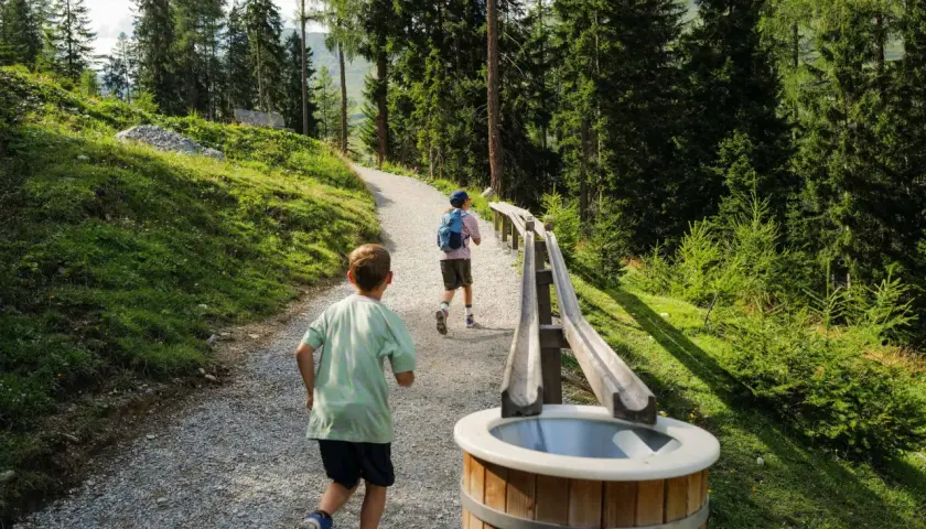 Zwei Kinder rennen auf einem Schotterweg an einer **outdoor kugelbahn** vorbei, umgeben von einem Wald und grünen Hängen.