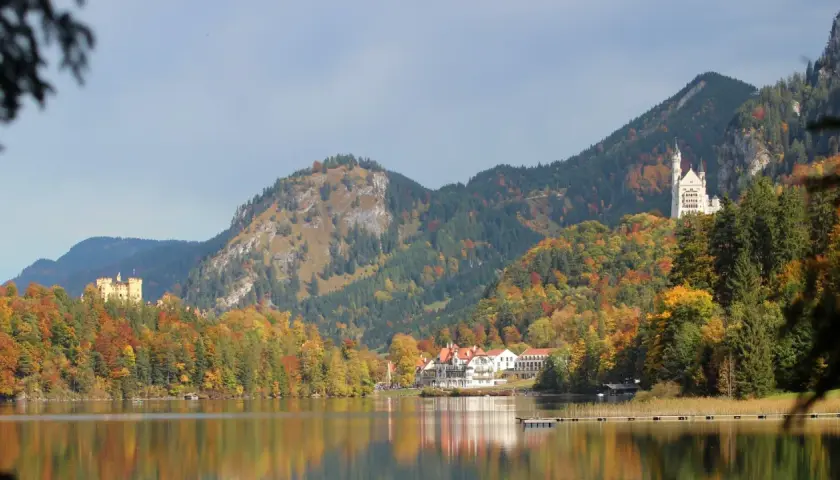 Zwei Märchenschlösser, Hohenschwangau und Neuschwanstein, thronen über einem See in Bayern, umgeben von bunten Herbstwäldern.