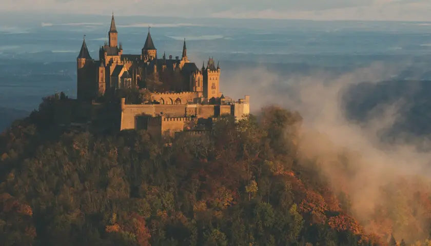 Burg Hohenzollern thront majestätisch auf einem Berg, umgeben von nebligen Herbstwäldern. Ein atemberaubender Aussichtspunkt.