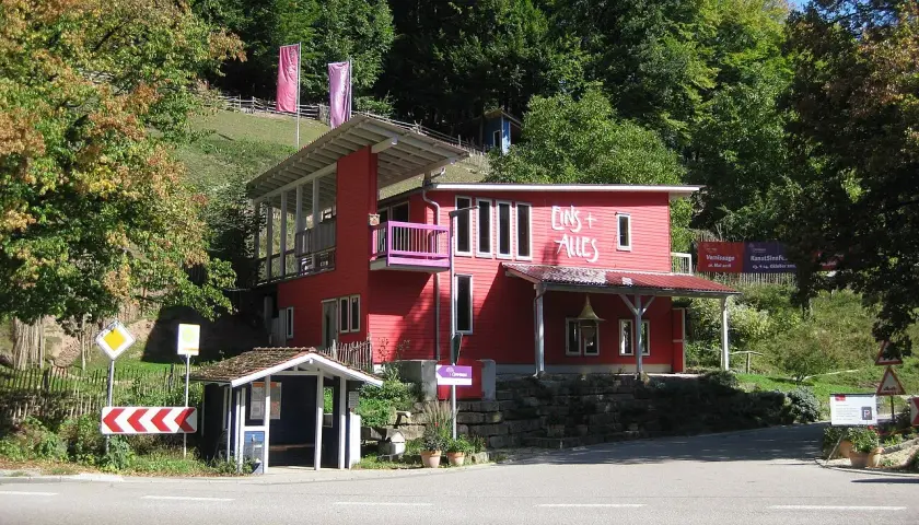 Rotes Haus mit der Aufschrift "Eins + Alles" am Waldrand. Ein kleiner Bushäuschen steht daneben.