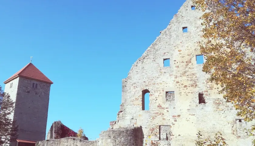 Ruinen der Lichtenburg Ostheim unter blauem Himmel. Ein Wehrturm mit rotem Dach ragt empor. Herbstlaub schmückt die alten Mauern.