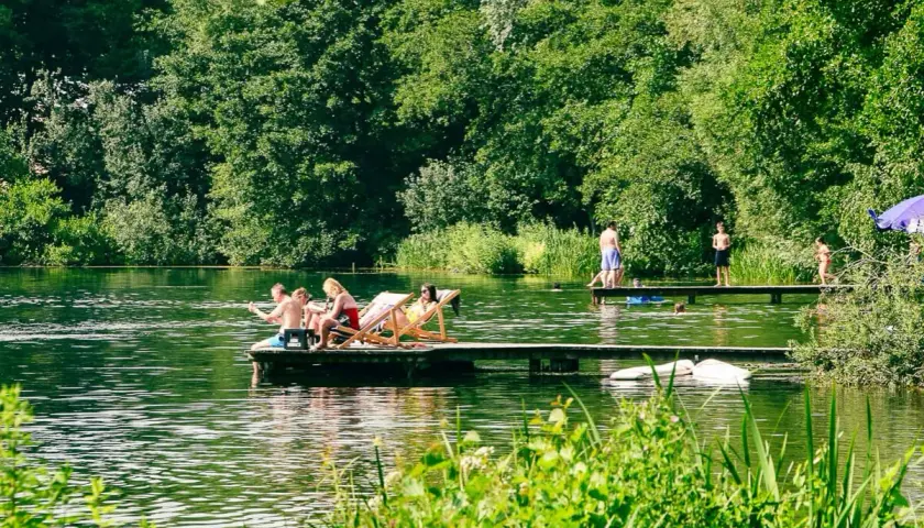 Menschen entspannen sich auf einem Floß und genießen die Sonne an einem Münchner See. Andere stehen auf einem Steg, einige baden im Wasser.