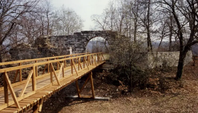 Holzbrücke führt zu einer steinernen Torbogenruine von Hofen, umgeben von kahlen Bäumen und Herbstlaub.
