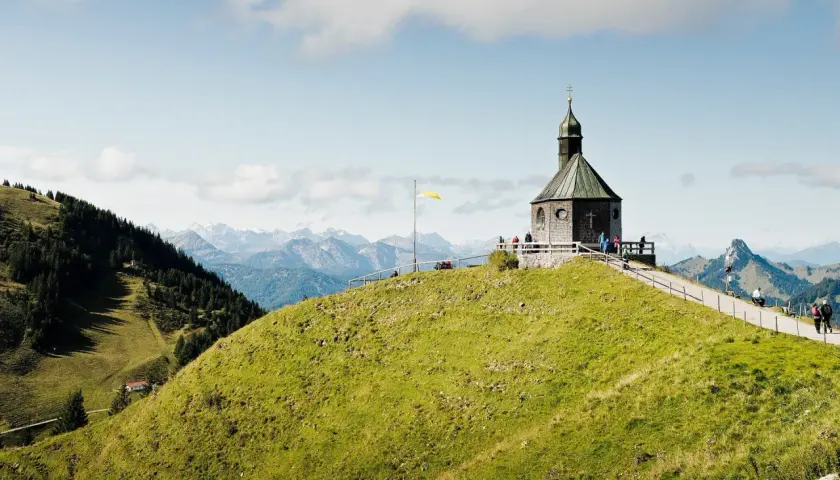 Blick auf das alte Wallberghaus auf einem grünen Hügel mit Bergpanorama. Eine Wanderung hierher lohnt sich für diese Aussicht.
