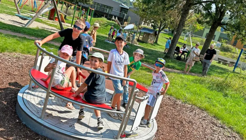 Kinder drehen sich auf einem Karussell auf dem Spielplatz. Ein Junge mit Sonnenbrille lacht, während er sich festhält. Der Spielplatz hat auch eine Schaukel und eine Rutsche, die an einen Flugzeugflügel erinnert.