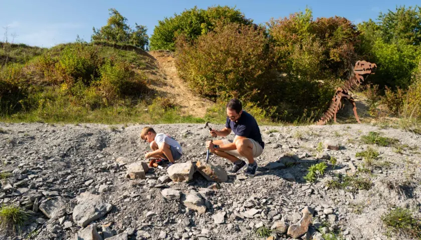 Ein Vater und sein Sohn graben nach Fossilien. Im Hintergrund steht ein Skelett eines Dinosauriers.