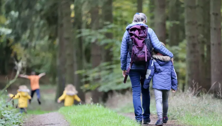 Familie genießt einen Waldspaziergang bei Aschaffenburg. Kinder in gelben Jacken rennen voraus, während eine Frau mit einem Kind Hand in Hand den Weg entlanggeht.