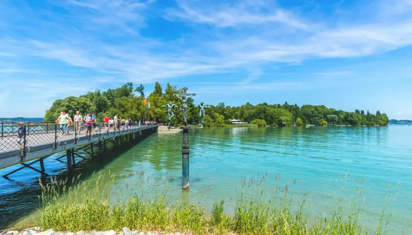 Menschen spazieren auf einer Brücke am Bodensee. Ein Denkmal mit Kreuzigungsgruppe steht im Wasser. Hier kann man in Konstanz viel unternehmen.