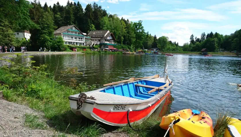 Ein Ruderboot namens "Irene" liegt am Ufer eines Sees im welzheimer Wald. Im Hintergrund ein Hotel und viele Menschen genießen den Tag.