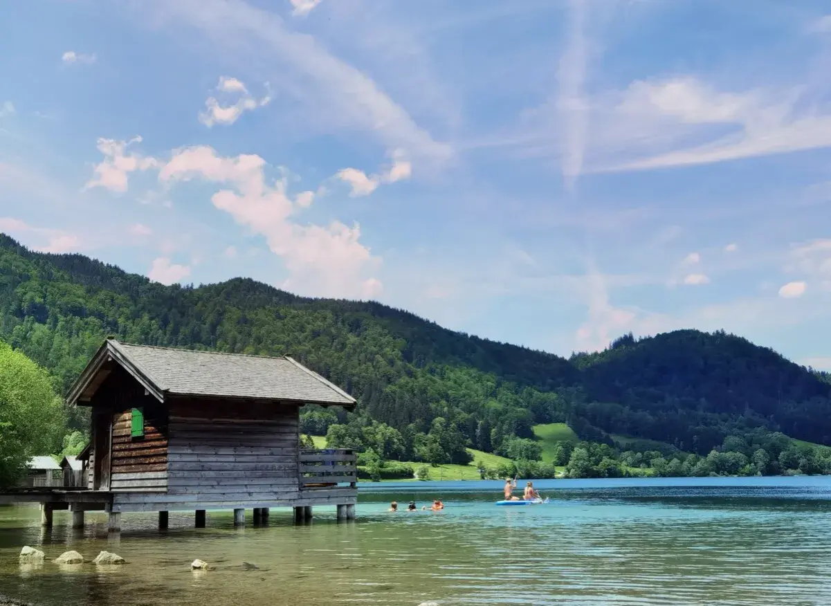 Holzhütte am See, Menschen planschen im Wasser. Perfekt für **Bayrischzell wandern mit Kindern** und Erfrischung.