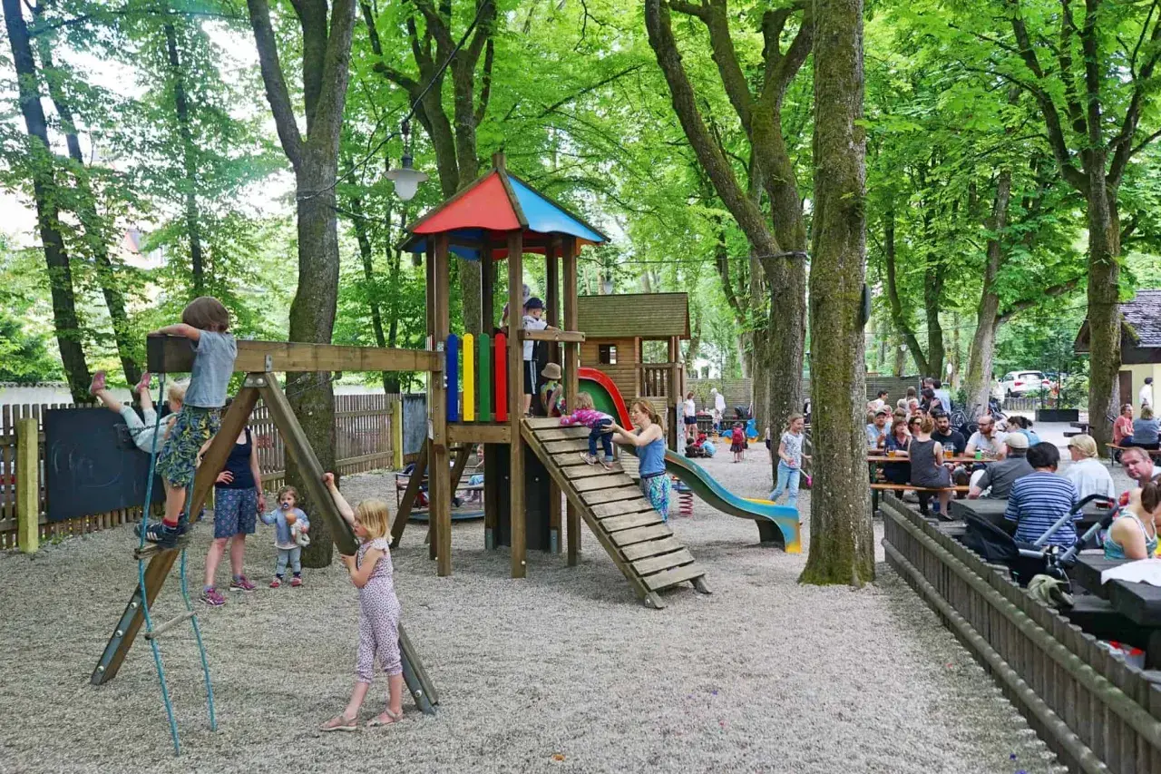 Kinder spielen im **Biergarten mit Spielplatz** unter schattigen Bäumen. Erwachsene sitzen an Tischen.