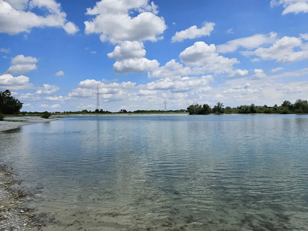 Ein sonniger Tag am Hollerner See mit blauem Himmel, weißen Wolken und klarem Wasser.