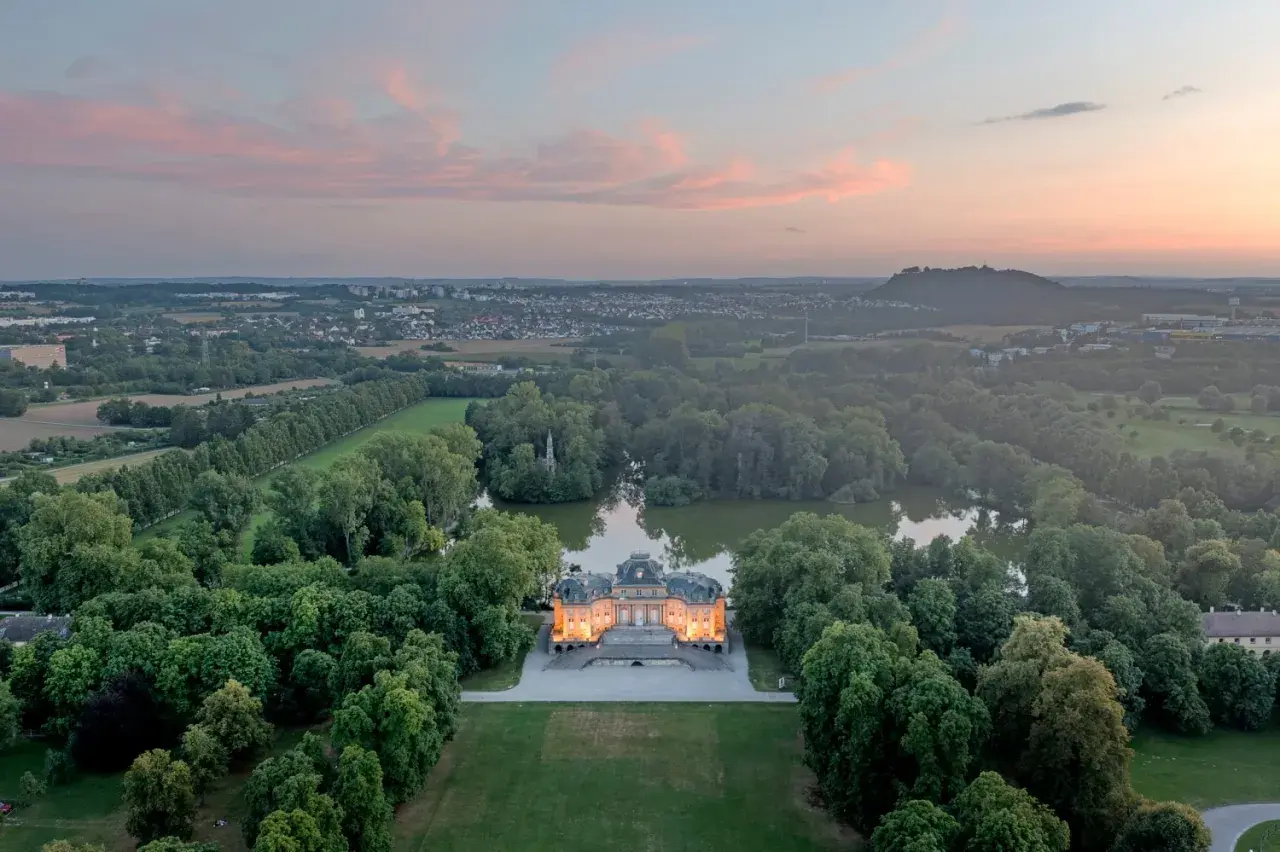 Schloss Monrepos Ludwigsburg, beleuchtet im Abendlicht, umgeben von einem See und üppigen Bäumen, mit Blick auf die Stadt.