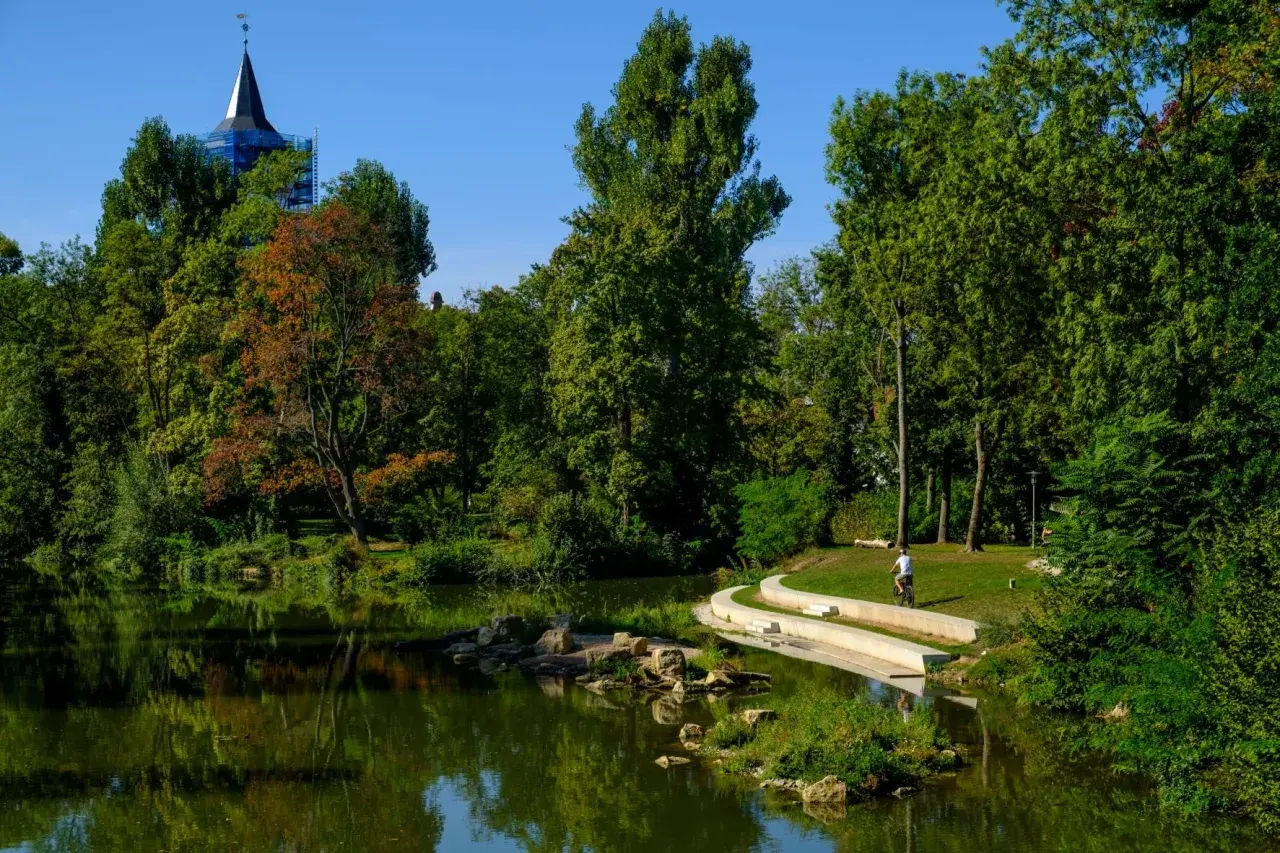 Ein Radfahrer fährt auf einem Weg am Wasser entlang, umgeben von Bäumen und einem Kirchturm.