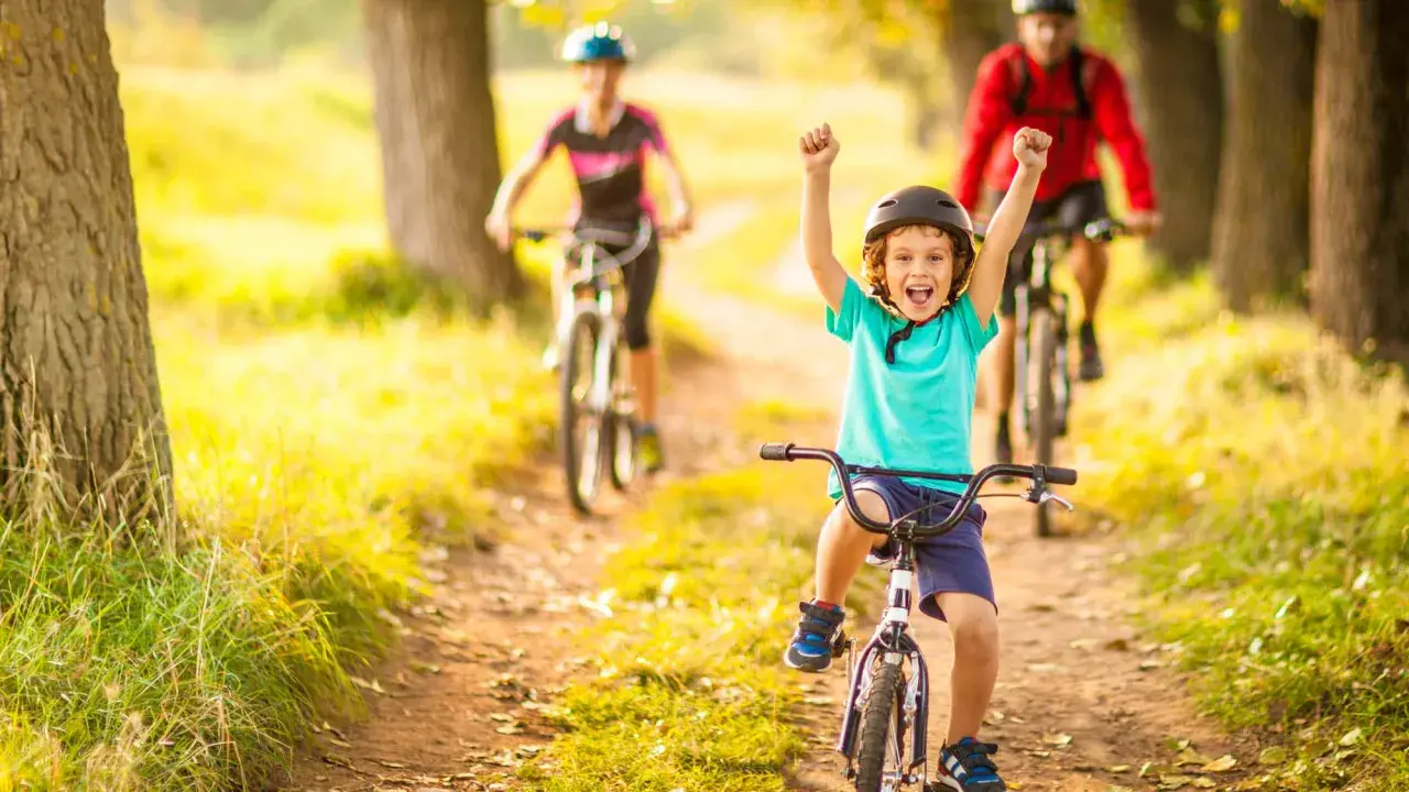 Glückliches Kind auf Fahrrad, feiert Erfolg bei leichten Radtouren mit Kindern. Eltern fahren im Hintergrund.