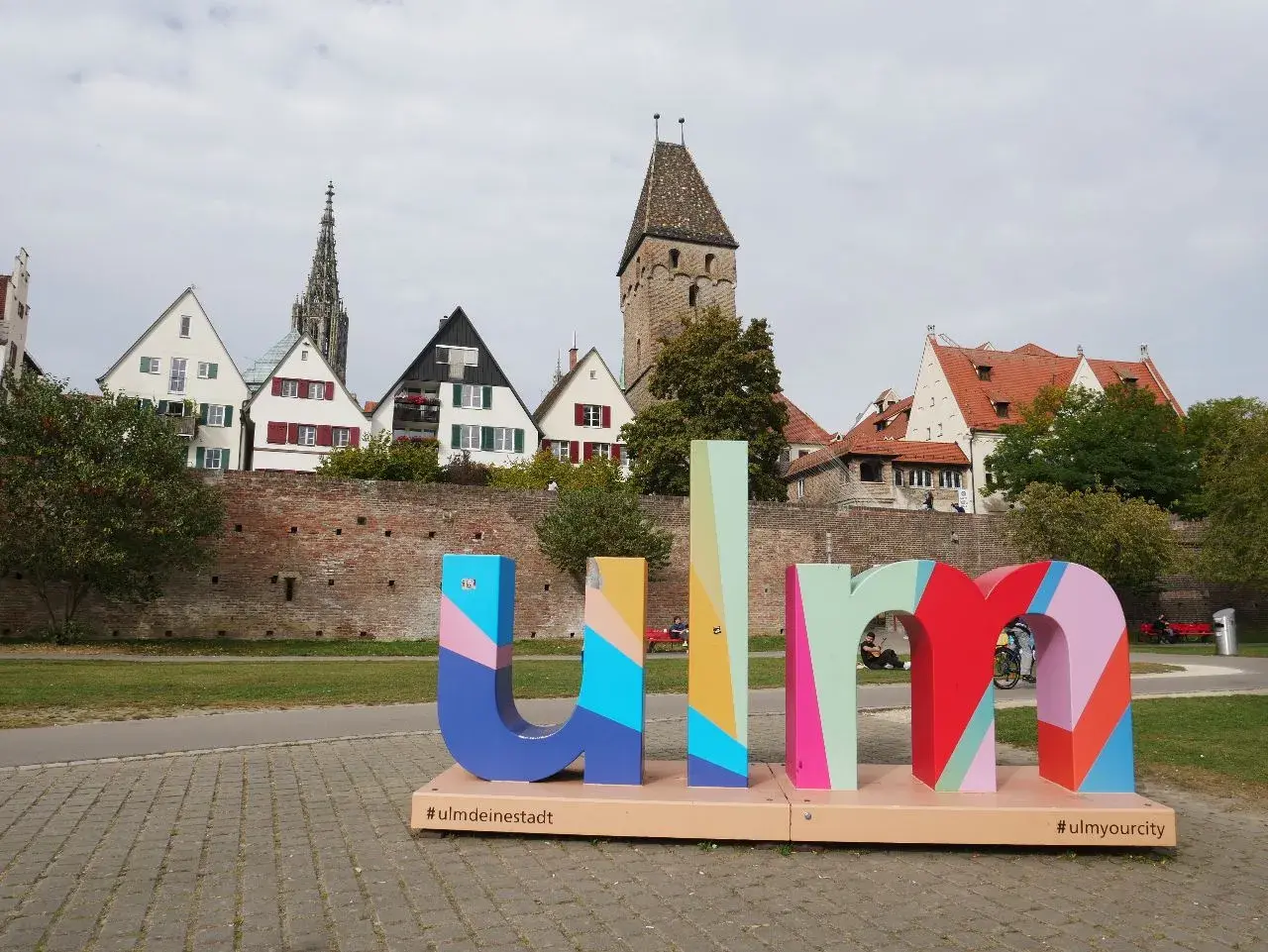 Bunte Buchstaben mit "#ulmdeinstadt" und "#ulmyourcity" vor historischer Kulisse. Hier kann man Ulm erleben!