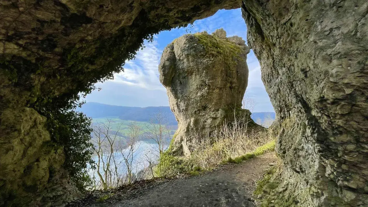 Blick durch eine Felsöffnung auf einen markanten Felsen mit Moosbewuchs. Ein Pfad führt entlang der Felsen. Eine tolle hohle Felsen Tour.