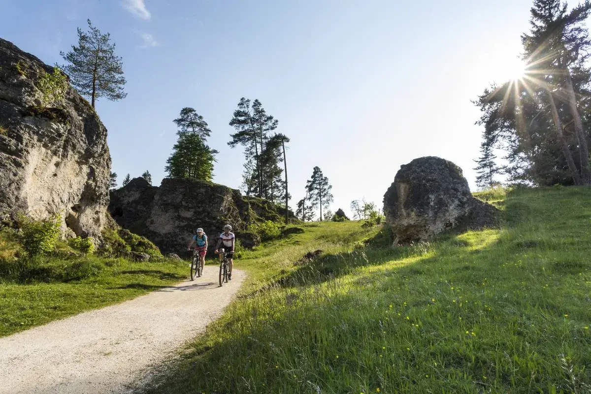 Zwei Radfahrer genießen eine Tour durch eine felsige Landschaft. Sie fahren auf einem Schotterweg, umgeben von grünen Wiesen und Bäumen.