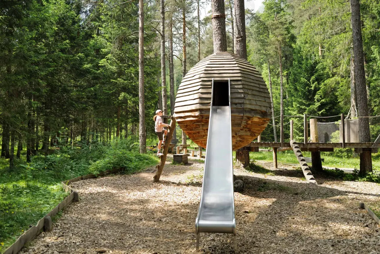 Ein Mädchen klettert auf einem Baumstamm an einem Waldspielplatz. Eine runde Holzhütte mit Rutsche steht im Mittelpunkt.