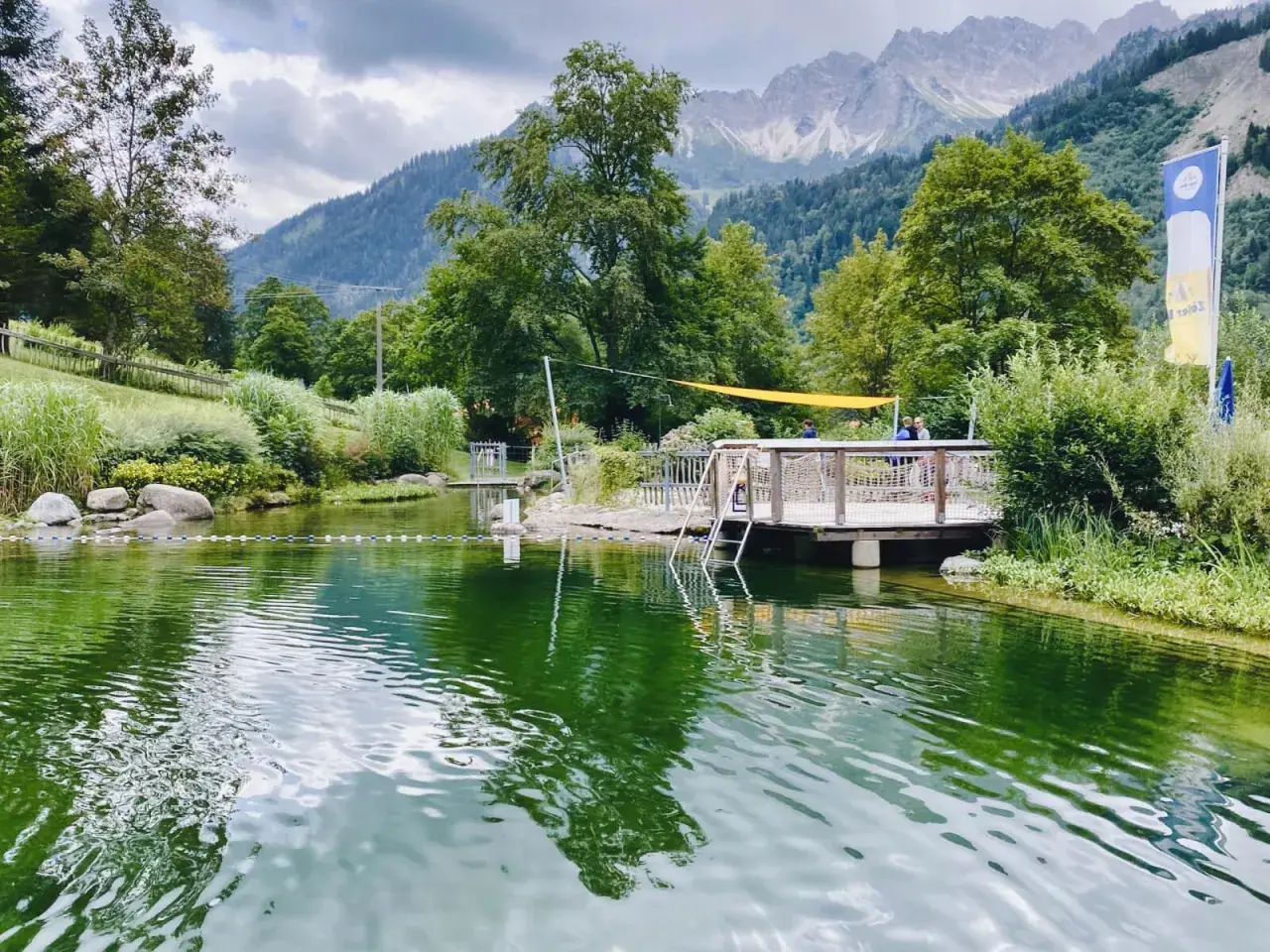 Ein idyllischer Badesee in Bad Hindelang, perfekt für das Wandern mit Kindern. Ein Steg mit Leiter lädt zum Sprung ins kühle Nass ein, während im Hintergrund majestätische Berge thronen.