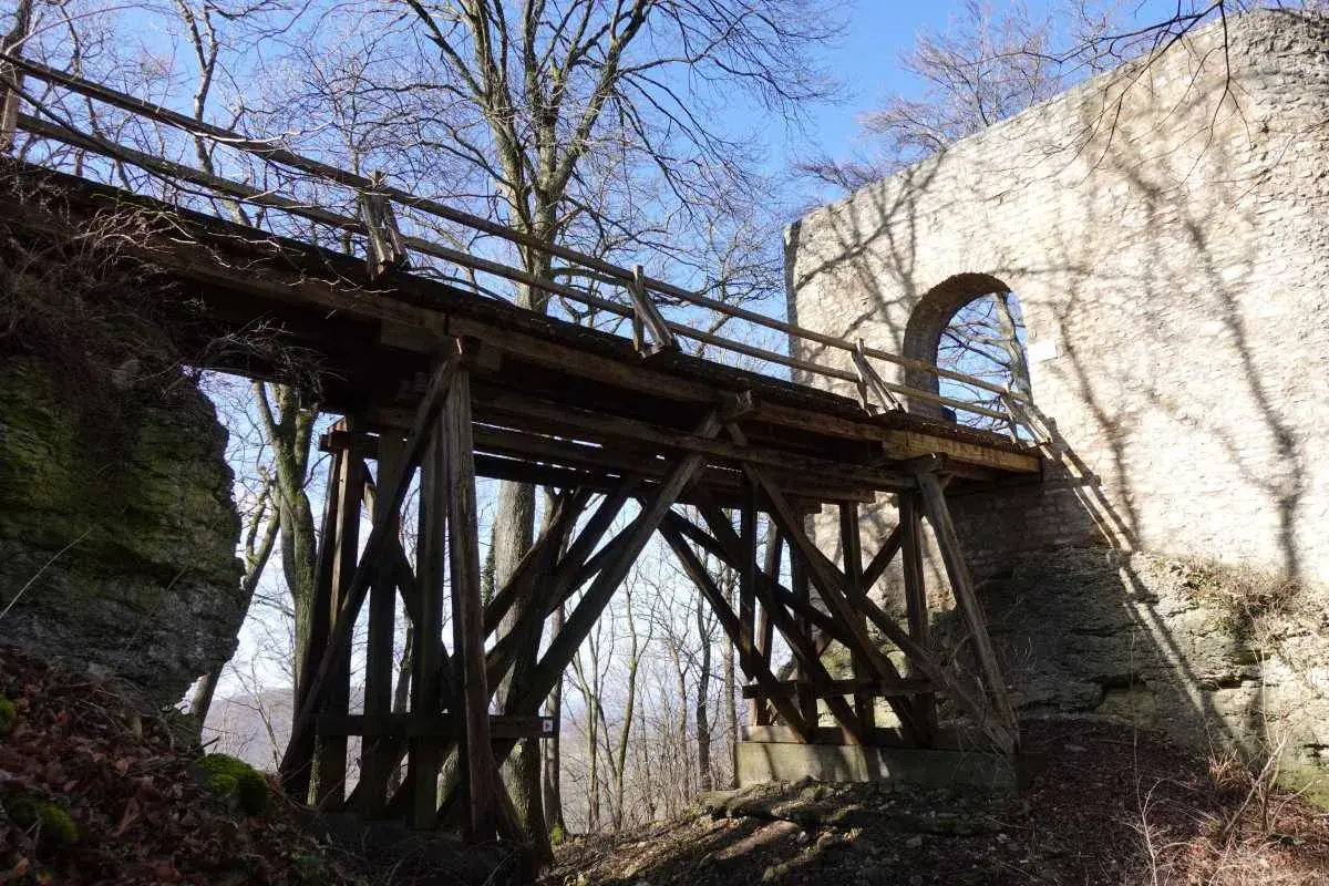 Holzbrücke führt zur Burgruine Rauber. Steinmauer mit Bogen und Schatten von Bäumen.