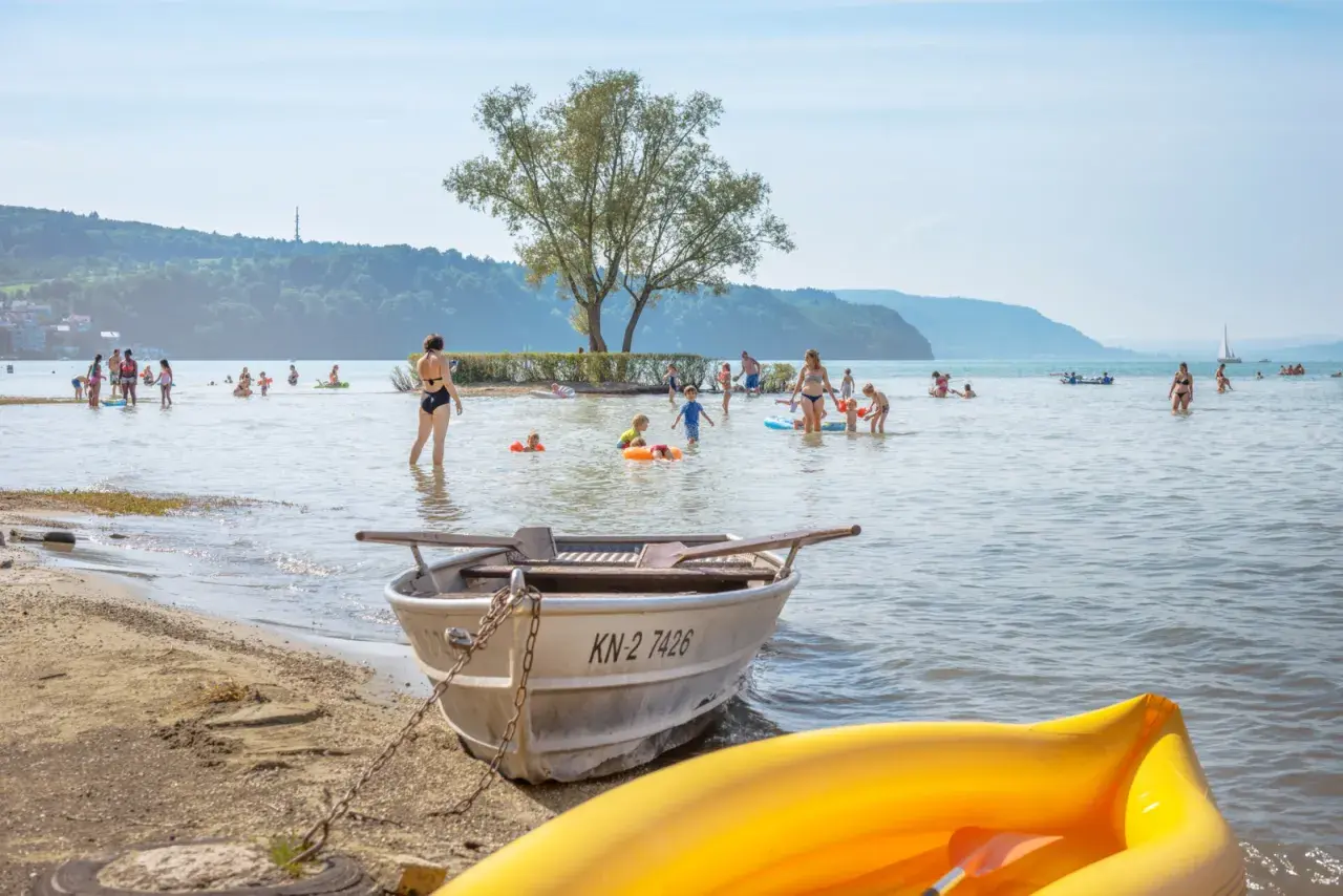 Menschen genießen das Bodensee Baden an einem sonnigen Tag. Kinder planschen im Wasser, während Boote am Ufer liegen.
