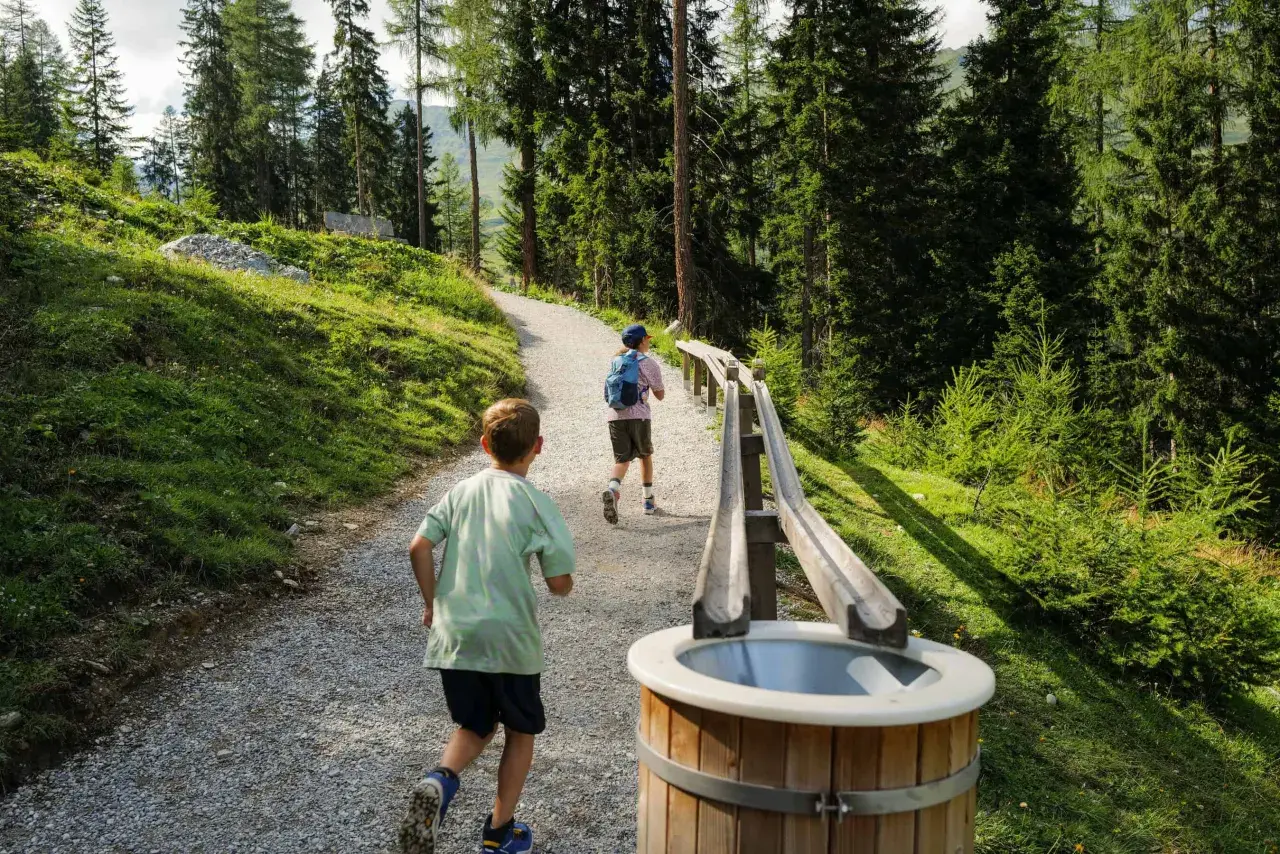 Zwei Kinder rennen auf einem Schotterweg an einer **outdoor kugelbahn** vorbei, umgeben von einem Wald und grünen Hängen.