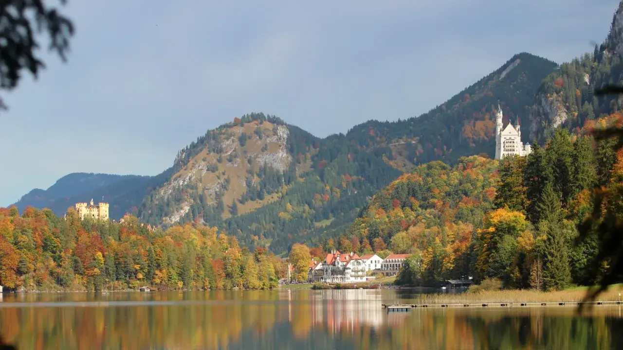 Zwei Märchenschlösser, Hohenschwangau und Neuschwanstein, thronen über einem See in Bayern, umgeben von bunten Herbstwäldern.