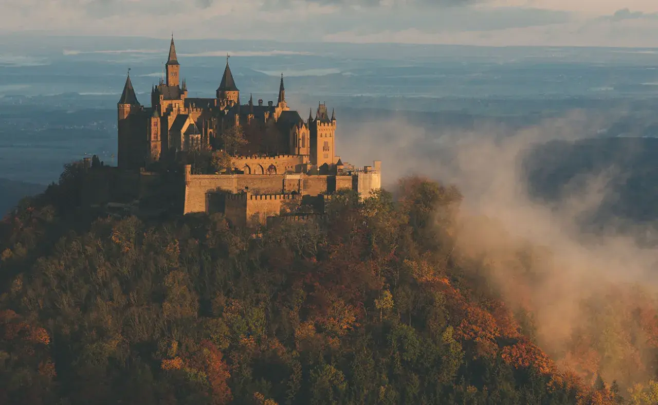 Burg Hohenzollern thront majestätisch auf einem Berg, umgeben von nebligen Herbstwäldern. Ein atemberaubender Aussichtspunkt.