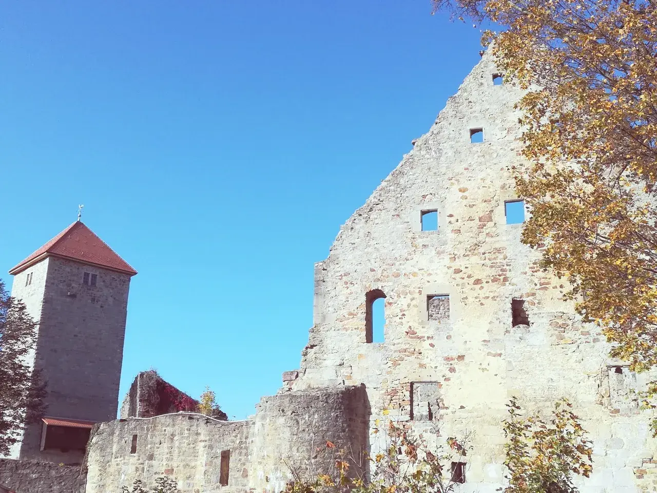 Ruinen der Lichtenburg Ostheim unter blauem Himmel. Ein Wehrturm mit rotem Dach ragt empor. Herbstlaub schmückt die alten Mauern.