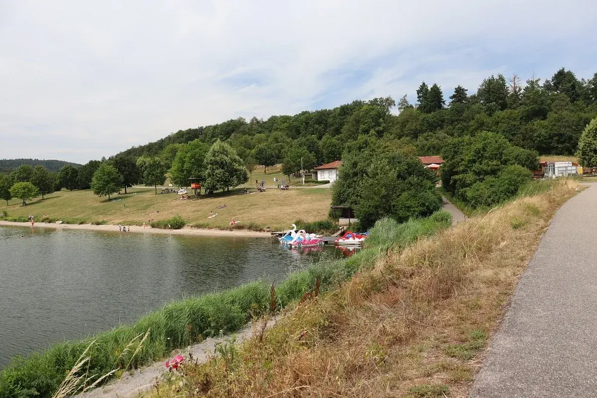 Sonniger Tag am badesee Zaberfeld. Menschen entspannen am Ufer, Kinder planschen im Wasser, Tretboote warten auf Ausflügler.