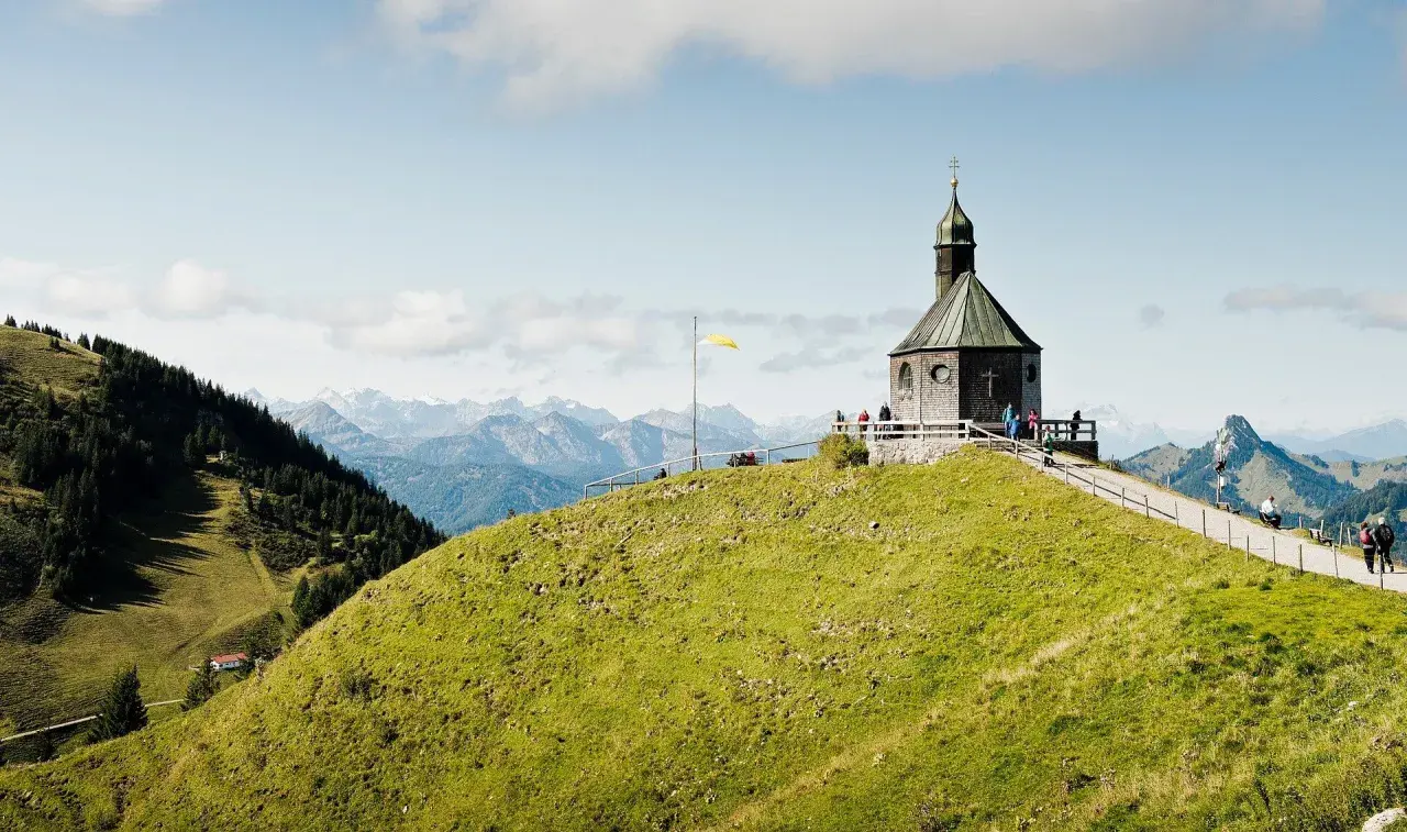Blick auf das alte Wallberghaus auf einem grünen Hügel mit Bergpanorama. Eine Wanderung hierher lohnt sich für diese Aussicht.