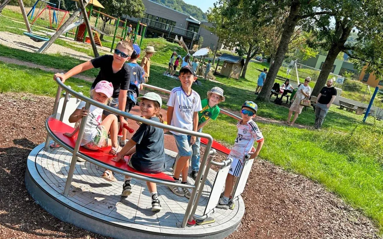 Kinder drehen sich auf einem Karussell auf dem Spielplatz. Ein Junge mit Sonnenbrille lacht, während er sich festhält. Der Spielplatz hat auch eine Schaukel und eine Rutsche, die an einen Flugzeugflügel erinnert.