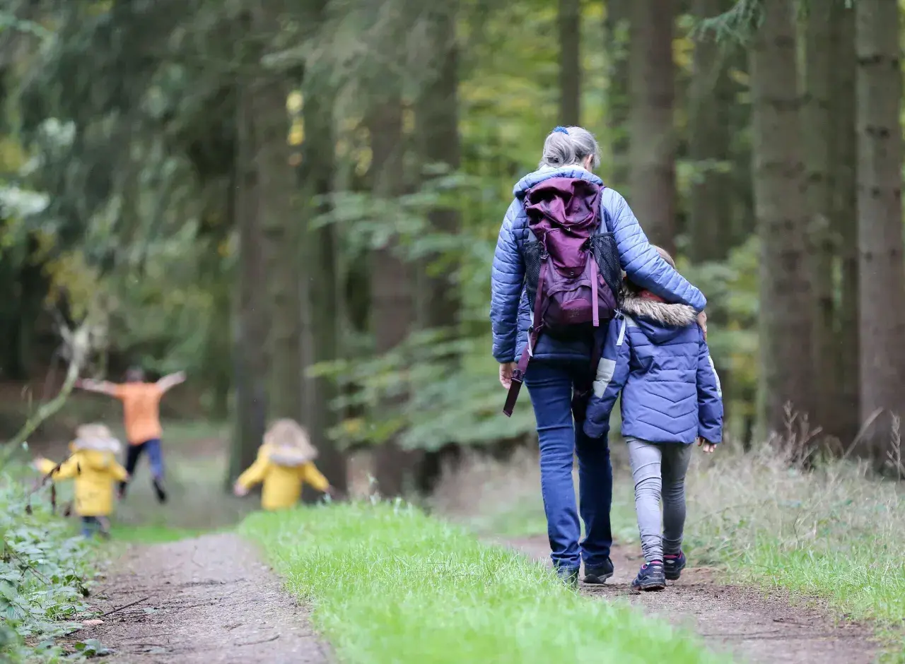 Familie genießt einen Waldspaziergang bei Aschaffenburg. Kinder in gelben Jacken rennen voraus, während eine Frau mit einem Kind Hand in Hand den Weg entlanggeht.