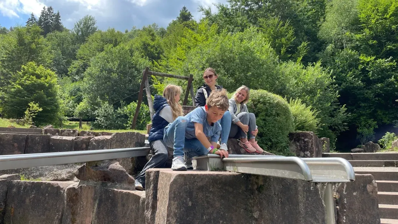 Familie genießt einen sonnigen Tag beim Wandern mit Kindern in der Pfalz. Ein Junge spielt am Wasserlauf, während die anderen entspannt zusehen.