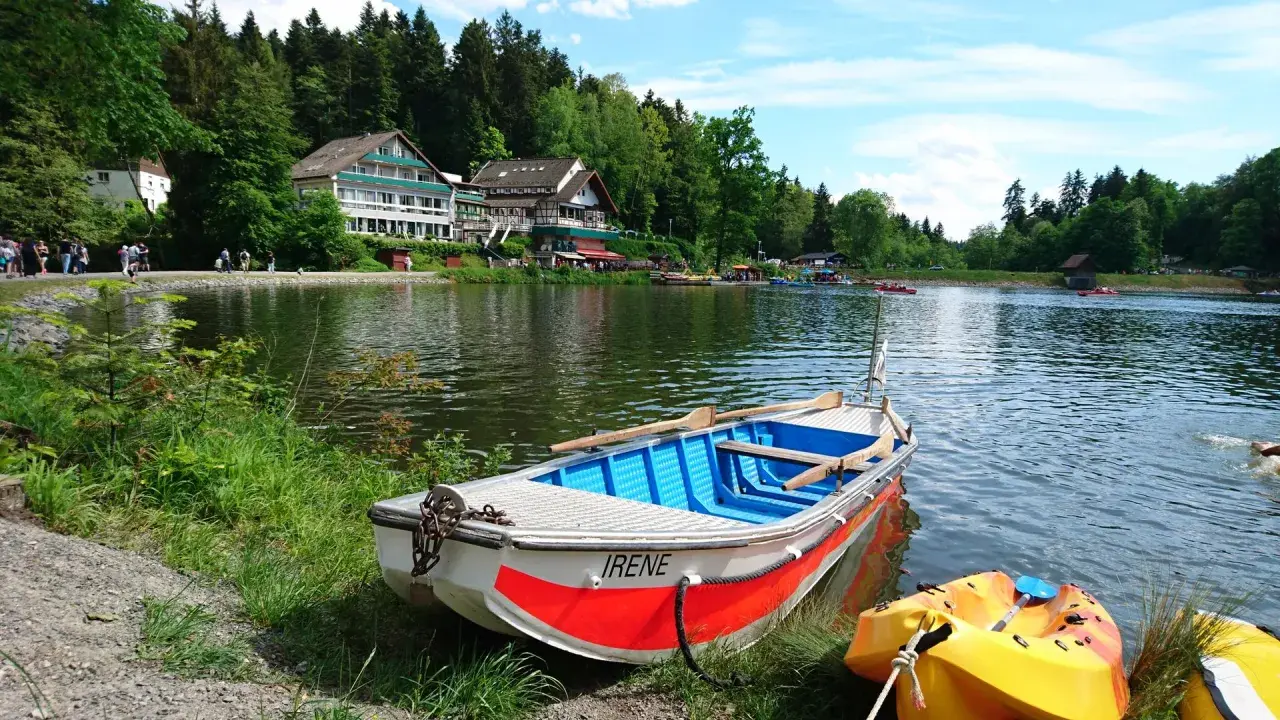 Ein Ruderboot namens "Irene" liegt am Ufer eines Sees im welzheimer Wald. Im Hintergrund ein Hotel und viele Menschen genießen den Tag.