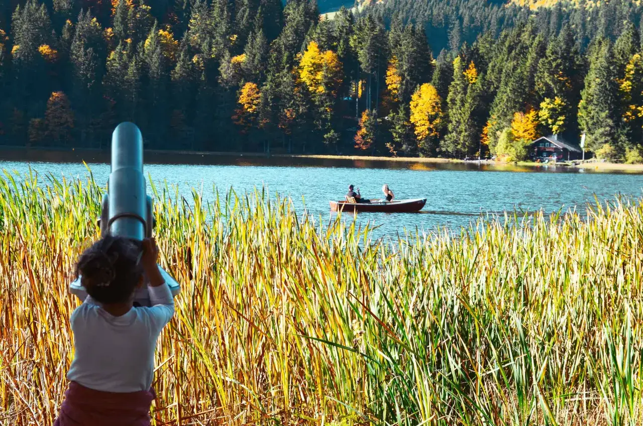 Ein Kind blickt durch ein Fernglas auf einen ruhigen See, umgeben von bunten Herbstwäldern. Ein Boot mit zwei Personen gleitet über das Wasser. Ein malerischer Anblick am Münchner See.