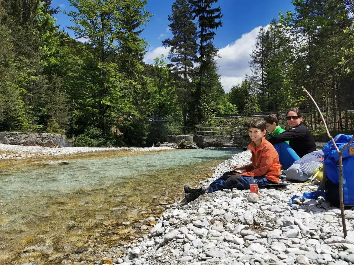 Familie genie&szlig;t Urlaub in Bayern am Fluss. Kinder und Mutter sitzen am Kiesstrand, umgeben von gr&uuml;nen W&auml;ldern.