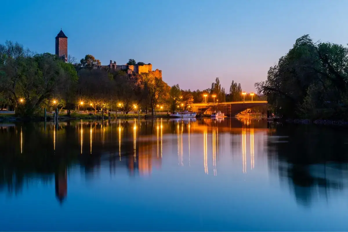Abendstimmung an der Saale: Burg Giebichenstein und beleuchtete Brücke spiegeln sich im Wasser. Ein perfekter Ort, um zu entspannen und zu sehen, was man in Halle machen kann.