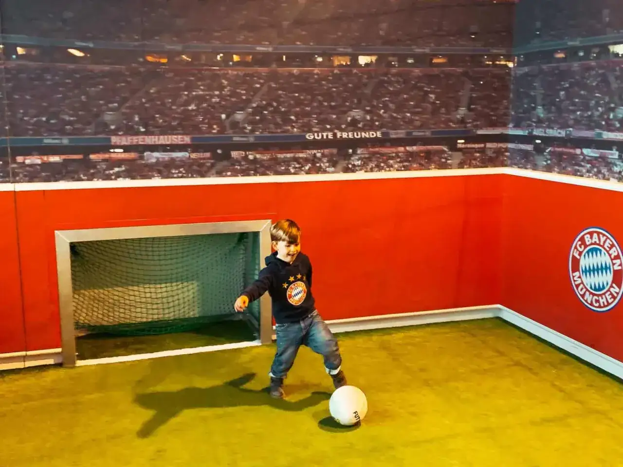 Kleiner Junge spielt Fußball in der Allianz Arena Spielwelt, vor einer Kulisse des Stadions.