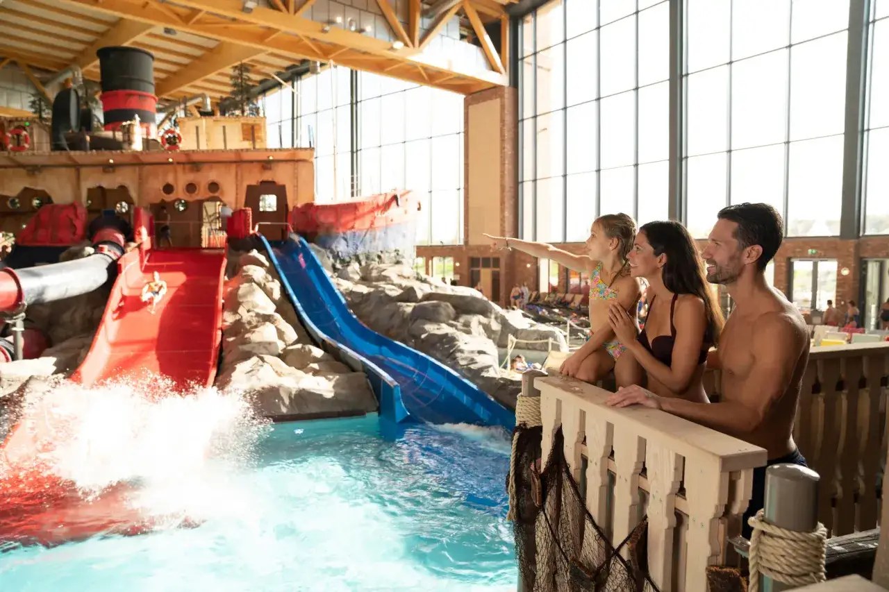 Familie beobachtet, wie Kinder im Schwimmbad die Rutschen hinunterfahren. Ein Junge saust die rote Rutsche hinunter, w&auml;hrend ein M&auml;dchen auf die blaue Rutsche zeigt.