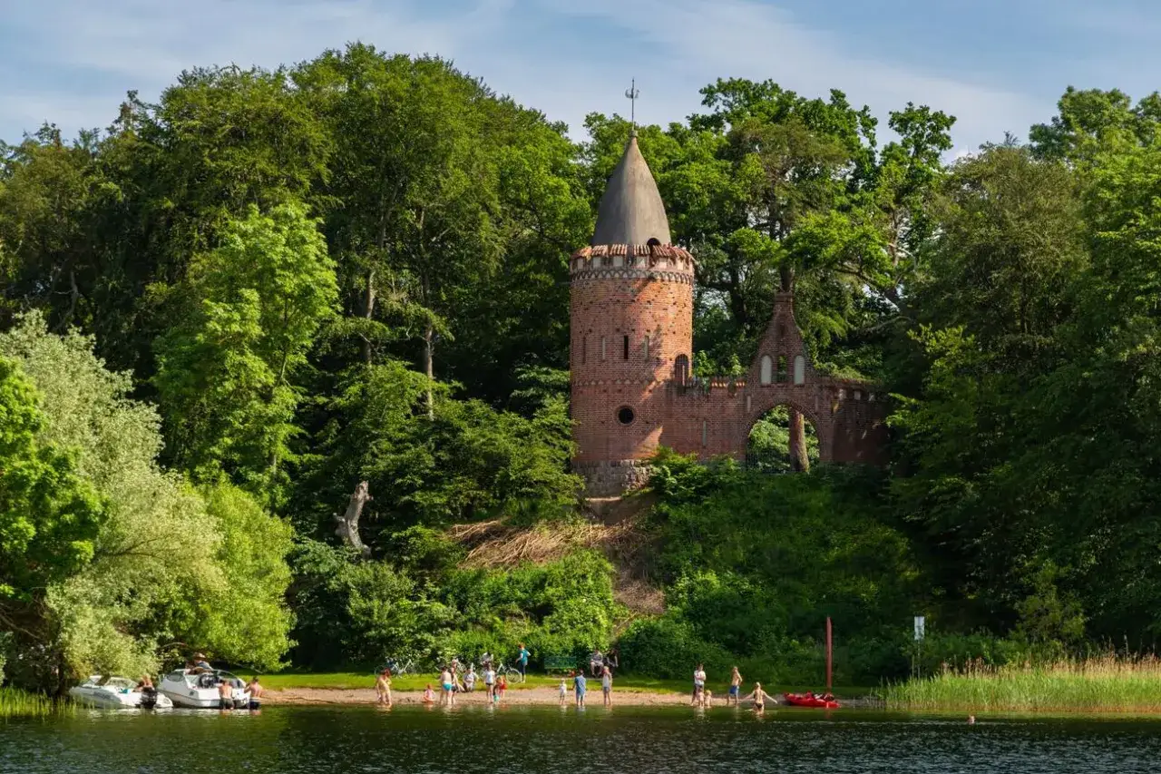 Ein versteckter Turm am Seeufer, ein Geheimtipp f&uuml;r Schwerin. Menschen baden und entspannen am Strand, umgeben von &uuml;ppigem Gr&uuml;n.