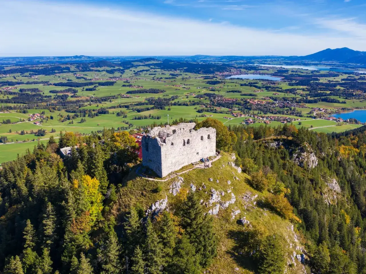 Ruinen der Burg Falkenstein bei Pfronten thronen auf einem Hügel, umgeben von Wäldern und mit Blick auf eine malerische Alpenlandschaft mit Seen.